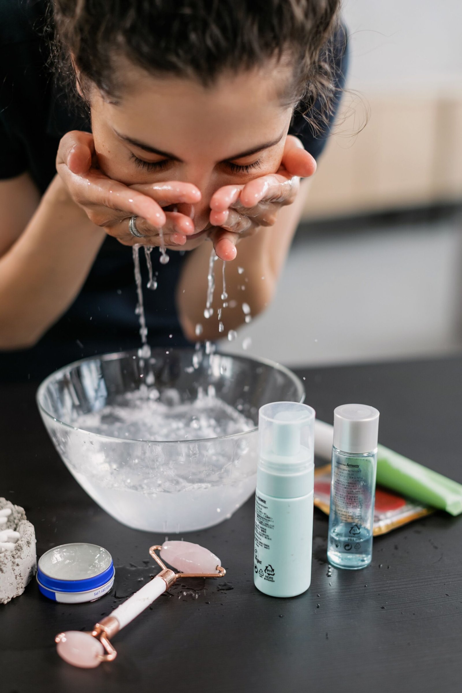 A woman washing her face with water over a bowl, surrounded by skincare products and a facial roller on a bathroom counter.