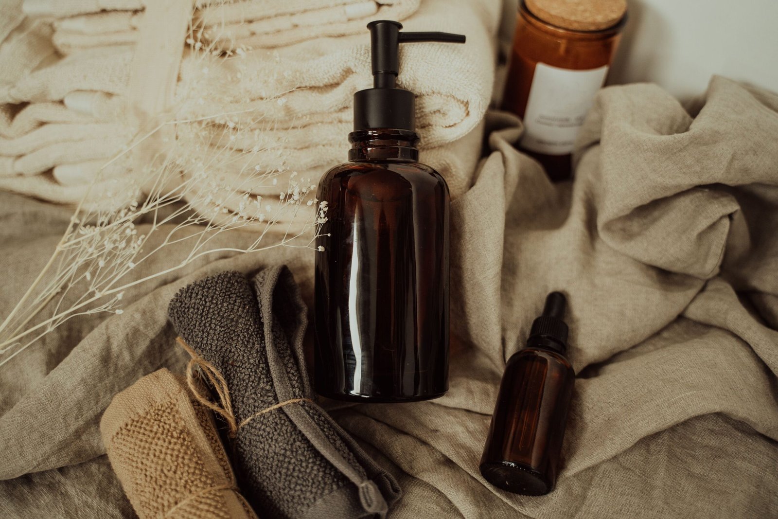 Amber glass skincare bottles arranged on beige towels with dried flowers in a neutral, minimalist bathroom setting.