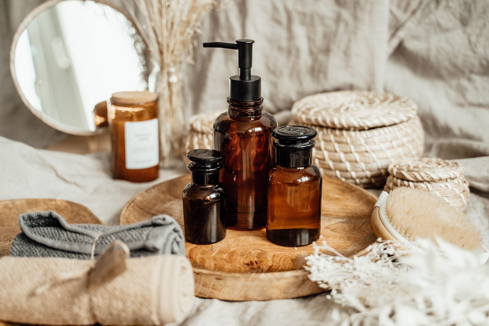 Amber glass cosmetic bottles displayed on a wooden tray with natural bath accessories, woven baskets, towels, and dried flowers.